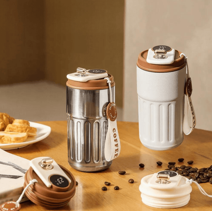 Two stylish thermal coffee mugs, one in stainless steel and the other in white, placed on a wooden table with coffee beans and pastries.