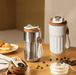 Two stylish thermal coffee mugs, one in stainless steel and the other in white, placed on a wooden table with coffee beans and pastries.