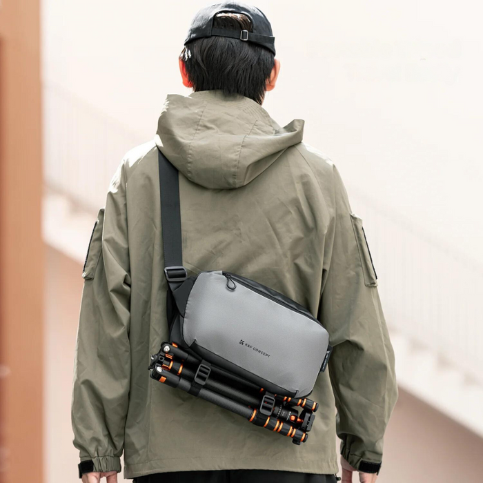 A person wearing a black cap and green jacket carries a gray K&F Concept camera sling bag with a tripod attached to the bottom.