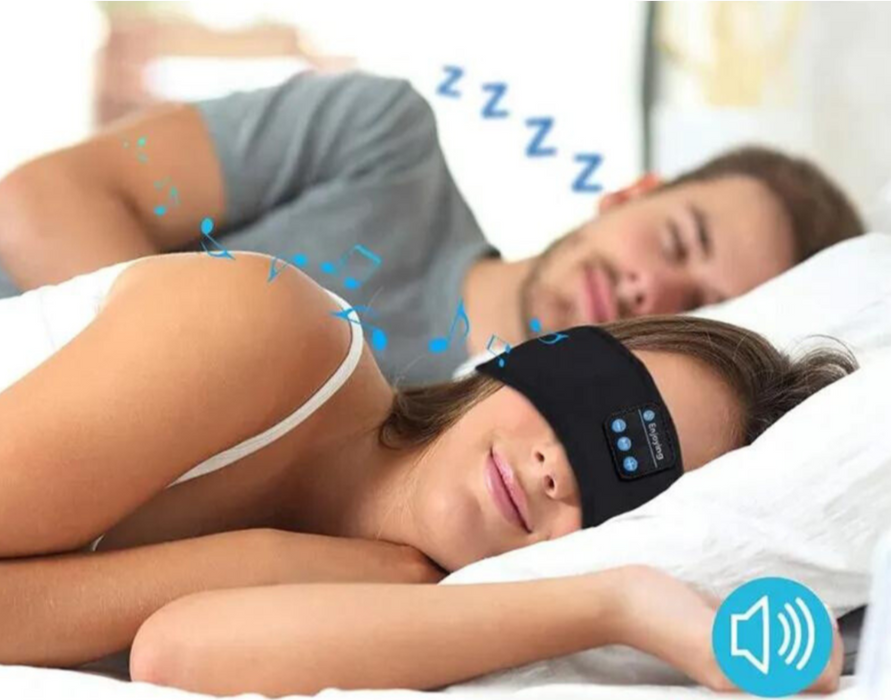 A woman and a man are lying in bed. The woman, in the foreground, is wearing a Bluetooth headband with musical notes depicted, while the man sleeps in the background.