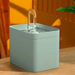 A green pet water fountain with a clear curved spout, set on a wooden surface against an orange background. The spout is positioned in the center of the fountain.