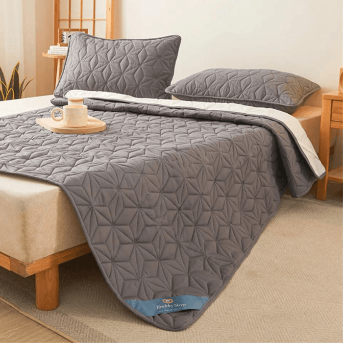 Gray quilted mattress cover pad folded back to show the white underside. Matching pillows are on the bed, with a cozy room setup including a small table, a plant, and a wooden tray with coffee mug on the bed.  