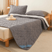 Gray quilted mattress cover pad folded back to show the white underside. Matching pillows are on the bed, with a cozy room setup including a small table, a plant, and a wooden tray with coffee mug on the bed.  