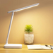 white desk lamp on a wooden desk, illuminating an open notebook and a pen. A glass vase with a green plant is also present, creating a warm, inviting workspace.