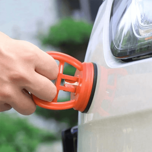 A close-up of a person’s hand attaching an orange suction cup to the curved surface of a car’s body, possibly for the purpose of dent removal or similar repair work.