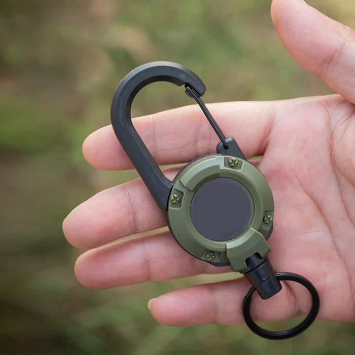 A close-up of a hand holding a compact retractable tool with a carabiner clip.