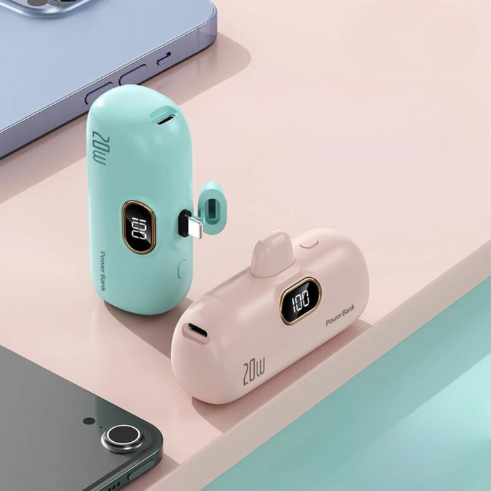 Two power banks (mint green and pink) with digital displays showing 100% charge, placed on a desk next to a smartphone.