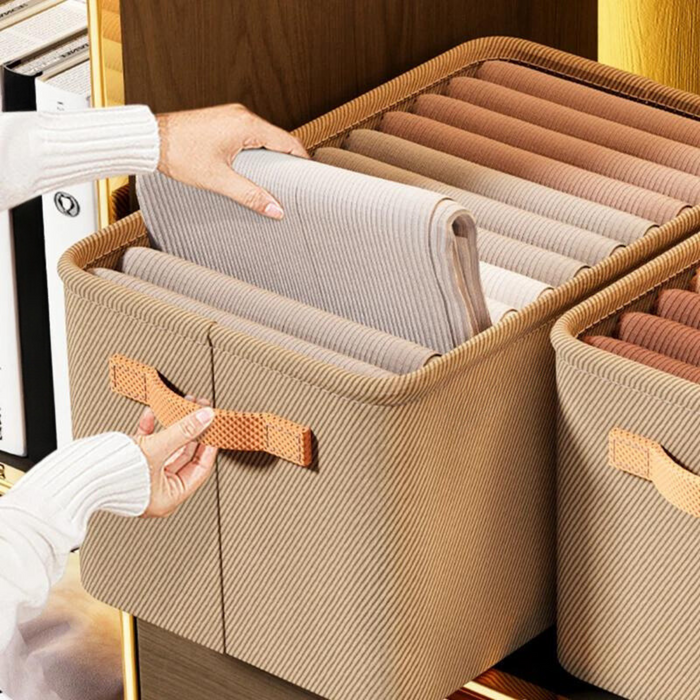 A single beige fabric storage bin with orange handles. The bin is filled with folded clothes arranged in rows while a woman's hands take one cloth from the bin. The label on the bin reads "Fashion." 