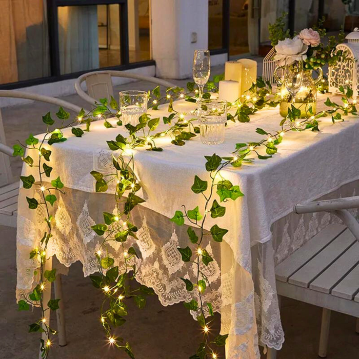 An elegantly decorated outdoor dining table covered with a white tablecloth and delicate lace details, beautifully lit by green ivy and fairy lights. This arrangement not only provides lighting but also adds a lush, natural touch to the setting, perfect for evening events.