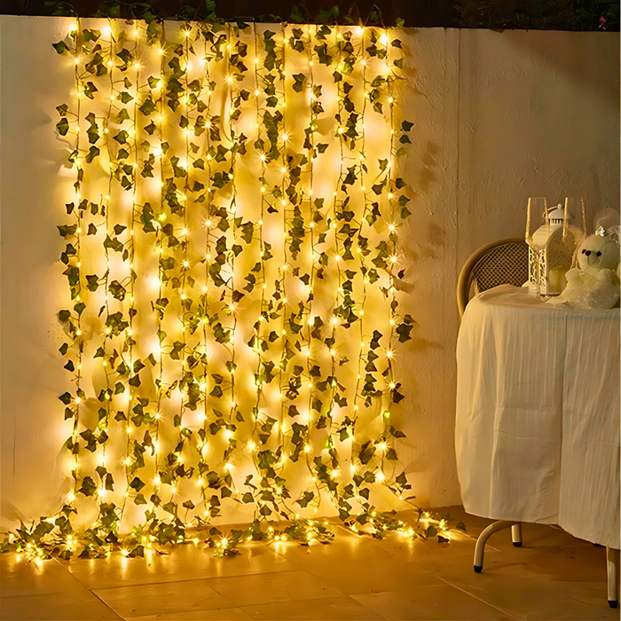 An outdoor setting illuminated by multiple strings of green ivy intertwined with small, bright lights hanging vertically against a white wall. This setup is part of a romantic dining area, complete with a table set for two, enhancing the atmosphere with its gentle, fairy-tale-like lighting.