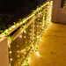 An outdoor balcony rail elegantly decorated with a string of green ivy leaves intertwined with warm white lights. The glowing lights provide a serene ambiance, enhancing the aesthetic appeal of the balcony, making it a perfect spot for evening relaxation.