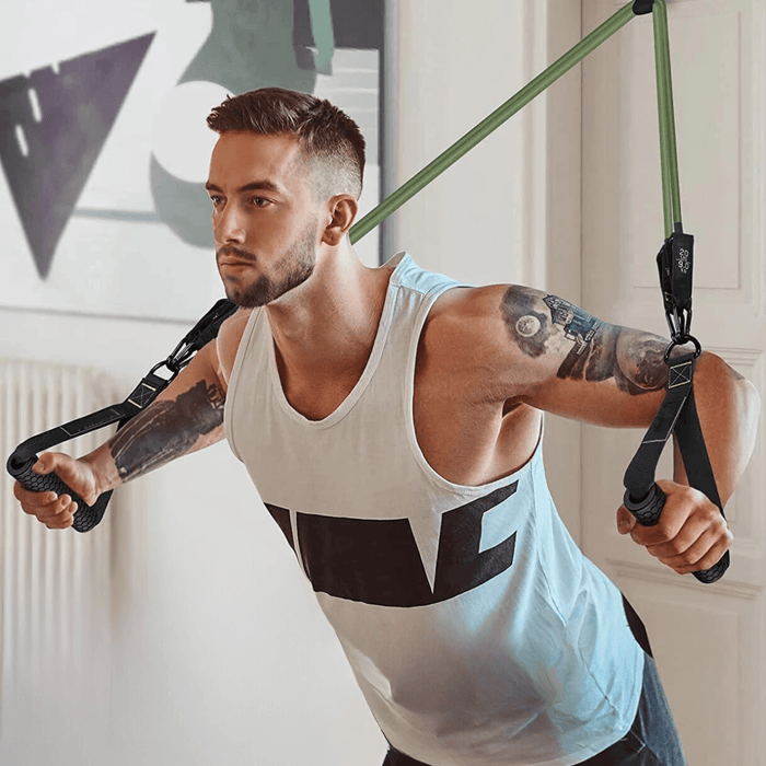 A young man with short dark hair and tattoos exercising with resistance bands indoors, wearing a white tank top.