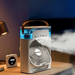 A portable air cooling fan with a blue top and brown handle on a bedside table, emitting mist. A digital clock shows "23:30" next to it.