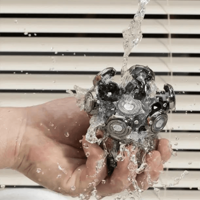 A hand holding a trimmer head under a stream of water. The trimmer head features multiple small circular blades, indicating it's waterproof and easy to clean.