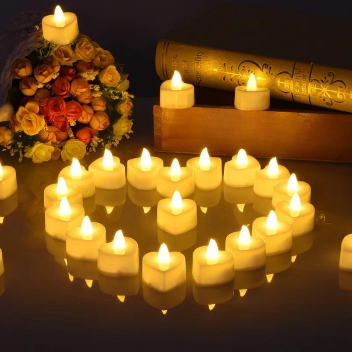 A warm, cozy setting with several white LED tea light candles arranged in a heart shape on a reflective surface. The candles emit a soft yellow glow, enhancing the romantic ambiance. Books and a bouquet of flowers are visible in the background.