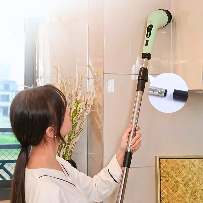 A woman using an electric cleaning brush with an extended handle to clean a high tile wall in a well-lit room with plants and a window in the background.