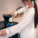 A woman using an electric cleaning brush to clean a stove top in a modern kitchen, with jars and cooking utensils visible in the background.