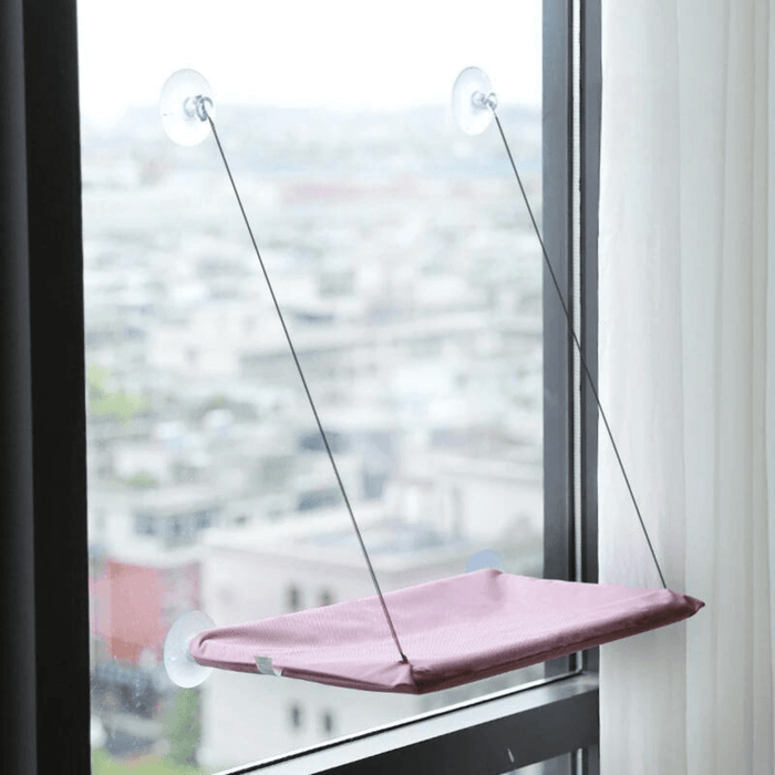 A pink version of the window-mounted cat hammock installed on a window, overlooking a cityscape.