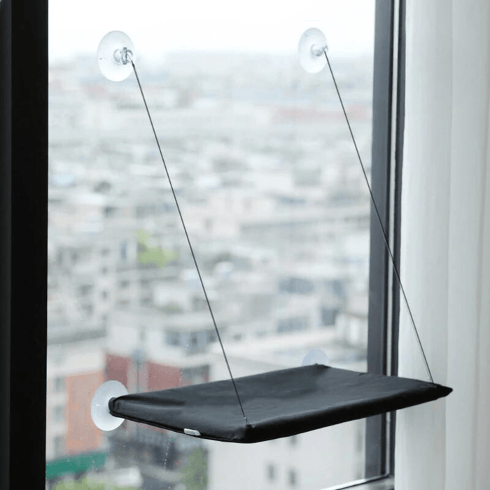 A black version of the window-mounted cat hammock installed on a window, overlooking a cityscape.