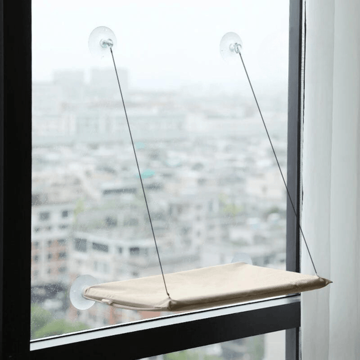 A khaki version of the window-mounted cat hammock installed on a window, overlooking a cityscape.