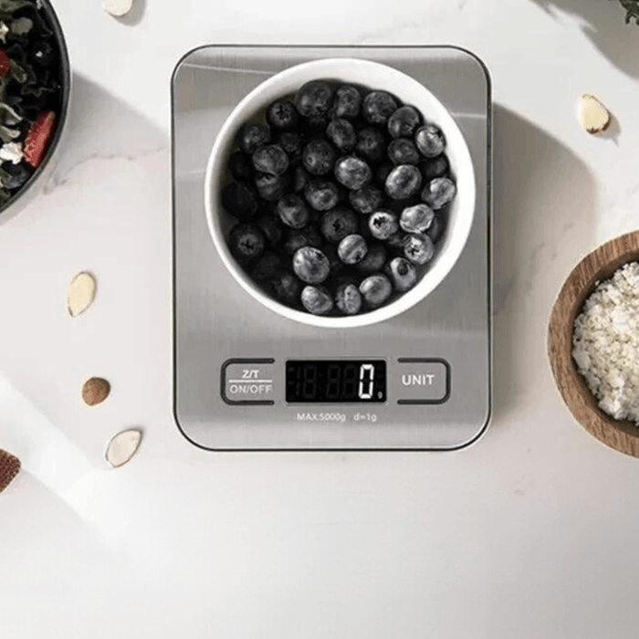 Stainless steel kitchen scale with a bowl of blueberries on top. On a white marble background.