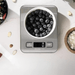 Stainless steel kitchen scale with a bowl of blueberries on top. On a white marble background.