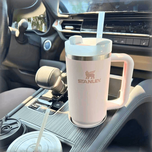 Stanley branded insulated travel mug with straw, prominently displayed in a car's cup holder. The mug is light pink with a logo, featuring a convenient handle for easy transport.