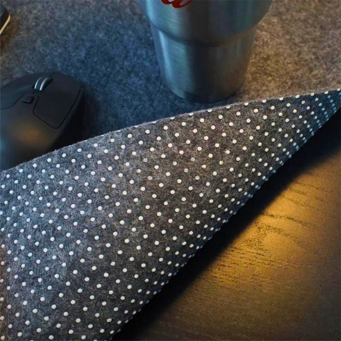 A close-up of a grey desk mat with a dotted non-slip backing, shown with a mouse and a metal tumbler on top, displayed on a dark wooden desk.