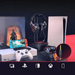 A gaming setup featuring various gaming devices including a Nintendo Switch, PlayStation 5, and an Xbox controller. A pair of black gaming headphones with RGB lighting is displayed on a stand. The background includes a game poster, and the foreground has icons representing compatibility with PC, PlayStation, Nintendo Switch, Xbox, and mobile devices.