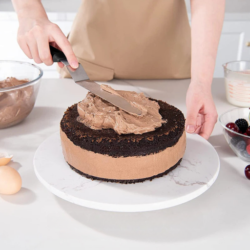 a person spreading chocolate frosting onto a chocolate cake using the spatula on a rotating cake stand.