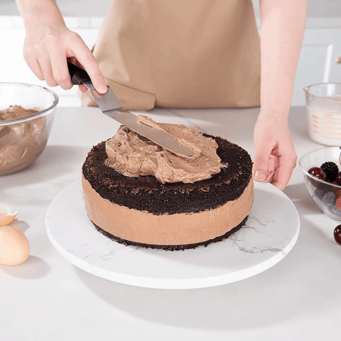 a person spreading chocolate frosting onto a chocolate cake using the spatula on a rotating cake stand.