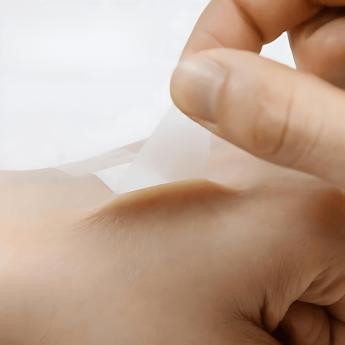 A close-up of a transparent adhesive bandage being peeled away from the back of the hand, demonstrating its flexibility and the ease with which it can be applied to the skin.