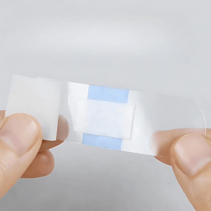 A transparent adhesive bandage being held between two fingers. This close-up reveals the thin and flexible nature of the bandage, designed to cover small wounds or cuts discreetly.