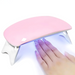 A hand placed under the pink nail dryer with blue LED light shining down, curing the nail polish. The pink dryer is positioned on its white legs, showcasing its function in action.