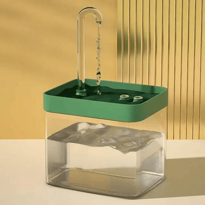 A pet water fountain with a green top and a clear base, filled with water and a clear curved spout extending from the top. It is set against a beige background.