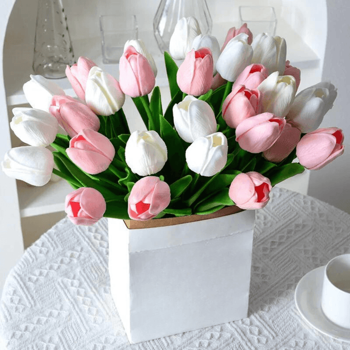 A bouquet of mixed tulips in shades of white, light pink, and baby pink with green leaves, arranged in a white box on a table.