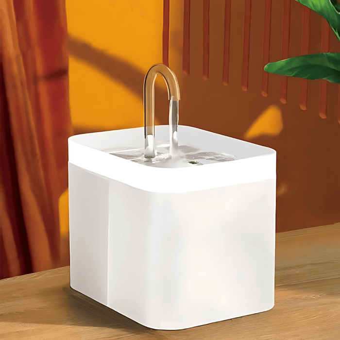 A white pet water fountain with a clear curved spout, set on a wooden surface against an orange background. The spout is positioned in the center of the fountain.