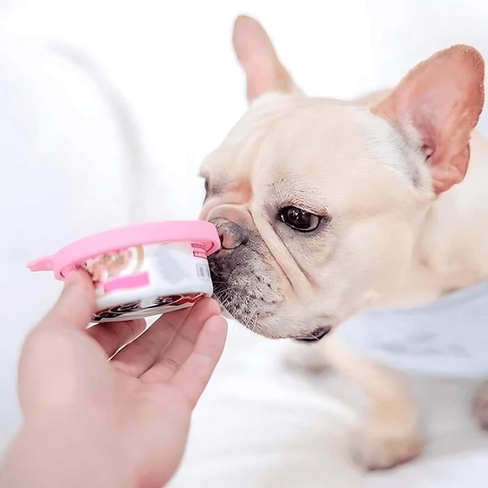 A French Bulldog sniffing a pink can lid as a human hand holds it open. This lid appears to be designed for pet food cans, providing a tight seal to keep the contents fresh.