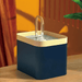 A dark blue pet water fountain with a light beige top, featuring a clear curved spout, placed on a wooden surface against an orange background.