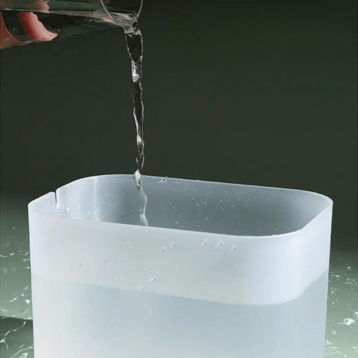 A clear plastic container is being filled with water from a glass. The container has a rounded rectangular shape and is partially filled with water.