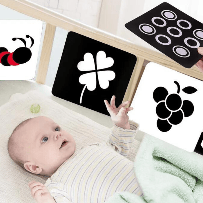 A baby lying in a crib looking at various "Baby Visual Stimulus Cards" propped up along the crib’s side, with designs including a black clover, a bunch of grapes, and circles.