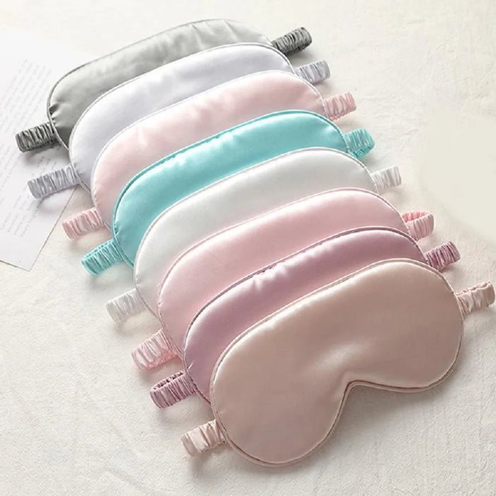 A selection of eight satin sleep masks in various pastel colors including pink, white, and blue, laid out in a row.