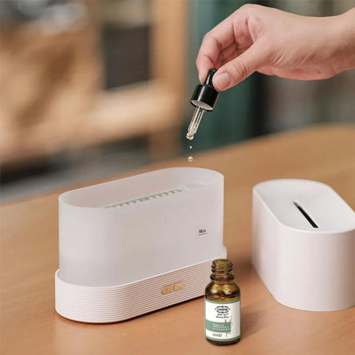 A white aroma diffuser with its top lid open. A hand is shown using a dropper to add essential oil. A bottle of essential oil is placed next to the diffuser.