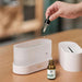 A white aroma diffuser with its top lid open. A hand is shown using a dropper to add essential oil. A bottle of essential oil is placed next to the diffuser.