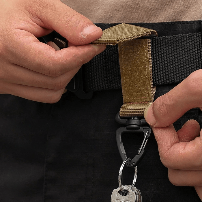 a tactical keychain strap being attached to a belt. The strap uses a Velcro closure to secure itself around the belt, with a carabiner clip at the end for holding keys or small tools.