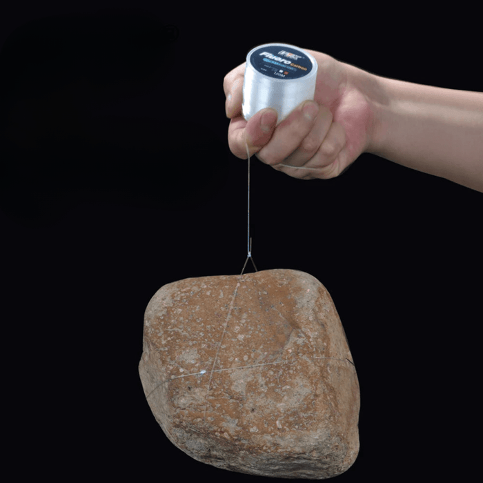 A hand holding a spool of fluorocarbon fishing line, suspending a large rock to demonstrate the line's strength and durability.