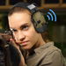 A woman aiming a rifle, wearing olive-green Arm Next earmuffs with an American flag patch. A blue sound wave icon indicates communication features ( Bluetooth).