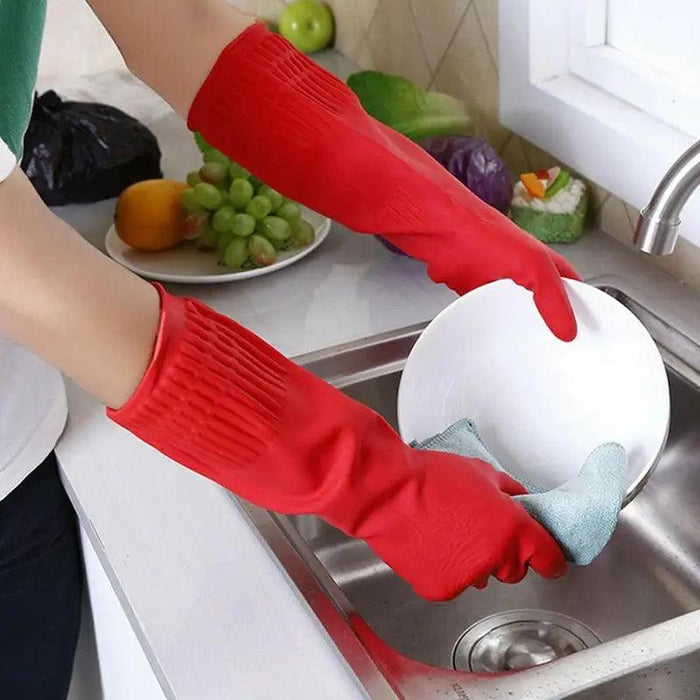 A person wears red cleaning gloves and holds a blue rag in his hand and cleans a plate over the sink with purple cabbage, lettuce, grapes, a lemon, a green apple and a blurred black garbage bag appearing in the background.