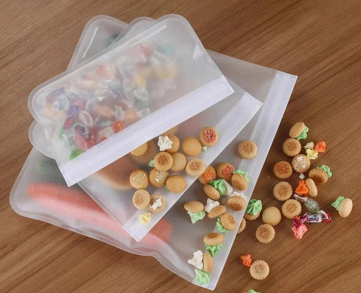 Reusable silicone food storage bags on a wooden surface, filled with various snacks and some spilled out.