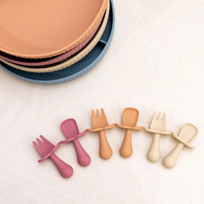 An assortment of baby utensils and silicone plates in various pastel colors, arranged on a white surface.